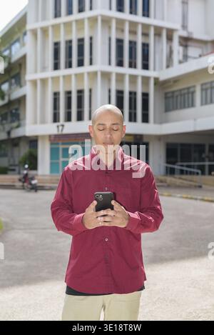 Glatzkopf italienischer Mann in burgunderrotem Hemd, der sein Smartphone überprüft, während er vor dem Universitätsgebäude bei hellem Sonnenlicht steht. Das Konzept des digitalen Kombi Stockfoto
