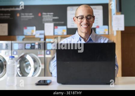 Glatzköpfiger männlicher Profi in blauem Button-Down-Hemd, der in der Wäscherei am Laptop arbeitet. Er setzt Technologie im Alltag ein. Echte Arbeit Stockfoto