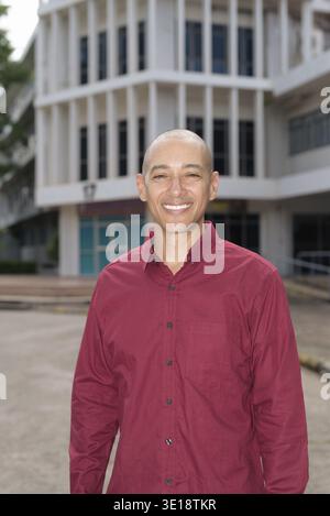 Stilvoller, kahlköpfiger italienischer Mann in burgunderfarbenem Button-Down-Hemd, der sich selbstbewusst vor dem modernen Universitätsgebäude an sonnigen Sommertagen posiert. Städtisch Stockfoto