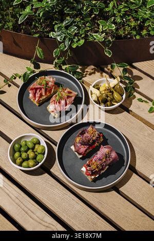 Auf zwei Tellern finden verschiedene Tapas mit Wurstwaren und Belägen sowie Schüsseln mit grünen und schwarzen Oliven in einer lebhaften Umgebung im Freien statt Stockfoto