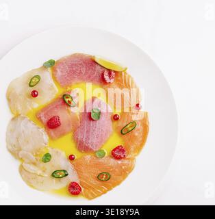 Eine künstlerische Anordnung verschiedener Fischscheiben, einschließlich Lachs, Thunfisch und Weißfisch, garniert mit Kräutern; Beeren und Zitrusfrüchte auf einem weißen Teller, Hi Stockfoto