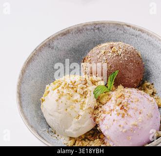 Drei köstliche Kugeln Eis in einer Schüssel umfassen Schokoladen-, Vanille- und Erdbeeraromen. Das Dessert wird mit zerdrückten Nüssen und einem Zweig belegt Stockfoto
