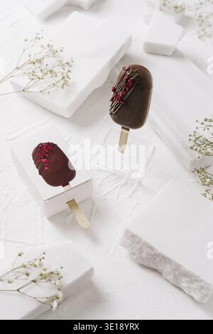 Zwei elegante Schokoladeneis ruhen auf einer strukturierten weißen Oberfläche. Sie sind mit lebhaften Belägen verziert und sorgen für eine verlockende Überraschung. Delicat Stockfoto