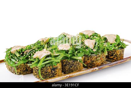 Farbenfrohe Sushi-Brötchen werden auf einem langen Teller serviert, der mit frischem Algensalat und einem Hauch cremiger Soße belegt wird. Ein Sprenkel Sesamsamen A Stockfoto