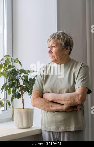 Seniorin steht am Fenster mit überkreuzten Armen, nachdenklich und nachdenklich im stillen Licht, reflektiert Einsamkeit und Erinnerung Stockfoto