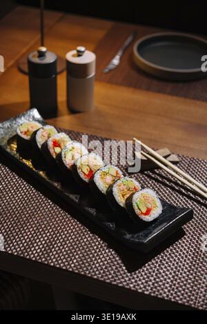 Exquisite Sushi-Brötchen serviert auf einem schwarzen Teller mit Gewürzen und Essstäbchen, bereit für ein Gourmet-Erlebnis Stockfoto