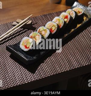 Exquisite Sushi-Brötchen serviert auf einem schwarzen Teller mit Gewürzen und Essstäbchen, bereit für ein Gourmet-Erlebnis Stockfoto