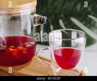 Lebendiger Hibiskustee, serviert in einer transparenten Teekanne mit passender Tasse auf einem Holztablett Stockfoto