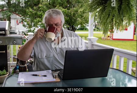 Ein Senior sitzt an einem Tisch auf einer Veranda und trinkt Kaffee aus einer großen Tasse, während er an einem Laptop arbeitet. Er hat Papiere und einen Stift in der Nähe. Die Einstellung zeigt einen grünen Rasen und einige Bäume. Stockfoto