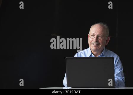 Grauhaariger Senior-Mann mit Schnurrbart, Brille und Laptop-Computer im Sitzen Stockfoto