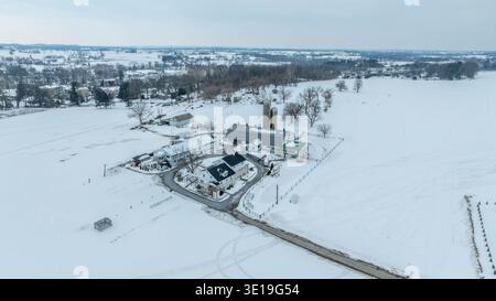 Ein Bauernhof ist im Winter mit dichtem Schnee bedeckt. Die Gebäude auf dem Bauernhof sind zusammen mit Feldern und Bäumen sichtbar. Die Aussicht zeigt eine ruhige Lage an einem bewölkten Tag. Stockfoto