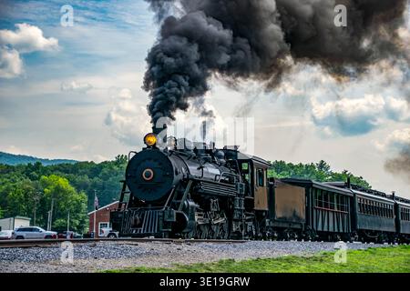 Eine Dampflokomotive fährt von einem Bahnhof weg. Dunkler Rauch steigt aus dem Motor auf, während Güterwagen hinter sich fahren. Autos stehen in der Nähe. Stockfoto