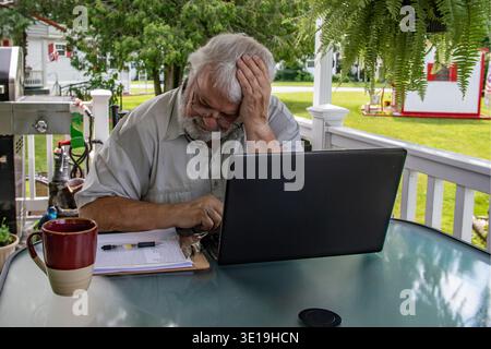 Ein Mann sitzt an einem Tisch im Garten und konzentriert sich auf seinen Laptop. Er sieht gestresst aus und hat eine Kaffeetasse neben sich. Bäume und ein Spielset sind im Hintergrund sichtbar. Stockfoto