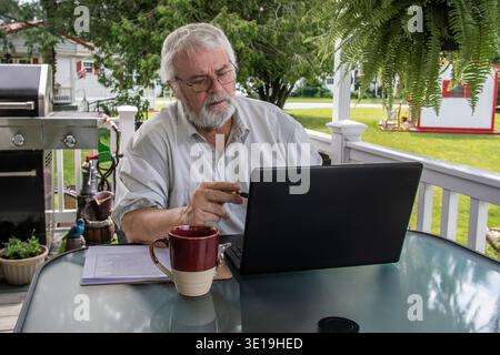 Ein Senior sitzt an einem Tisch auf einer Veranda. Er arbeitet an einem Laptop, während er einen Stift hält. Eine Kaffeetasse und Papiere liegen auf dem Tisch. Bäume und ein Grill sind an klaren Tagen im Hintergrund zu sehen. Stockfoto