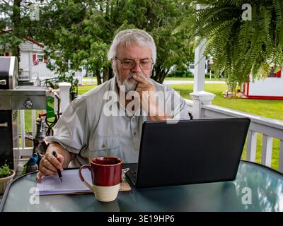 Ein Senior sitzt an einem Tisch auf einer Veranda und konzentriert sich auf seinen Laptop. Er hat eine Tasse Kaffee neben sich und schreibt in ein Notizbuch. Die Szene umfasst üppiges Grün und ein Haus im Hintergrund. Stockfoto