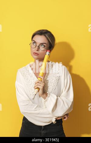 Nachdenkliche junge Frau, die eine Brille trägt, die großen Bleistift auf gelbem Hintergrund hält Stockfoto