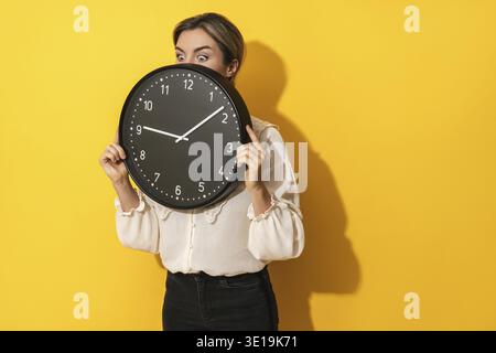 Eine junge Geschäftsfrau, die hinter einer großen Uhr auf gelbem Hintergrund rausblickt Stockfoto