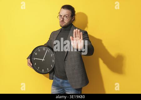 Ernsthafter Mann, der eine Brille trägt, die Uhr hält und Stoppgeste auf gelbem Hintergrund zeigt Stockfoto