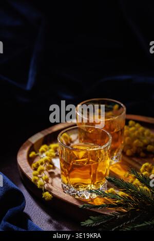 Zwei Gläser Whiskey oder Brandy mit Mimosa-Blumen auf Holztablett, Frühlingsgetränk-Konzept Stockfoto