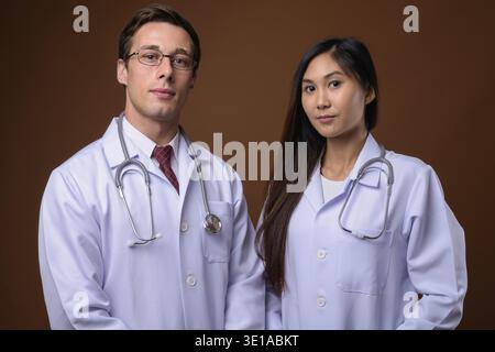 Studio shot Junger stattlicher Mann Arzt und junge schöne asiatische Frau Doktor gemeinsam gegen braunen Hintergrund Stockfoto