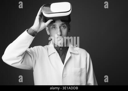 Studio Aufnahme von jungen schönen Mann Arzt tragen virtuelle Realität Headset vor grauem Hintergrund in Schwarz und Weiß Stockfoto