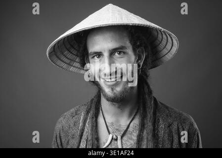 Studio-Aufnahme eines jungen gutaussehenden hispanischen Mannes mit Dreadlocks, der einen konischen Hut im asiatischen Stil auf grauem Hintergrund in Schwarz-weiß trägt Stockfoto