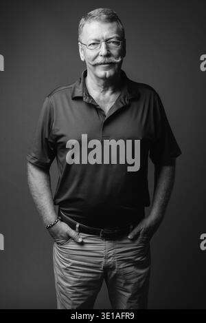 Studio-Aufnahme eines gutaussehenden älteren Mannes mit Schnurrbart, der eine Brille vor grauem Hintergrund in Schwarz-weiß trägt Stockfoto