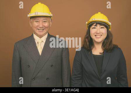 Studio mit einem Foto von einem reifen asiatischen Geschäftsmann und einer reifen asiatischen Geschäftsfrau, die vor braunem Hintergrund einen Hut trägt Stockfoto