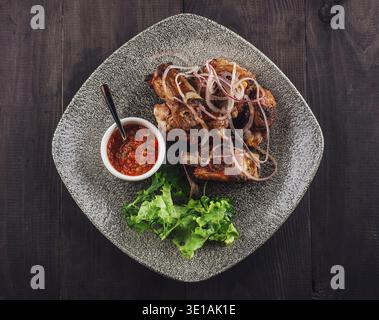 Gegrillte Schweinerippchen mit roten Zwiebeln und Kräutern, serviert auf einem Teller mit Sauce und frischem Salat auf dunklem Holzhintergrund Stockfoto