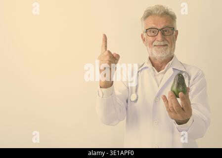 Studio geschossen von Stattlichen älteren bärtigen Mann Arzt holding Avocado während Zeigefinger auf weißem Hintergrund Stockfoto