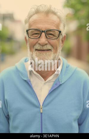 Angesichts der glücklich aussehenden älteren bärtigen Mann lächelnd beim Tragen blaue Jacke und Brille im Freien Stockfoto