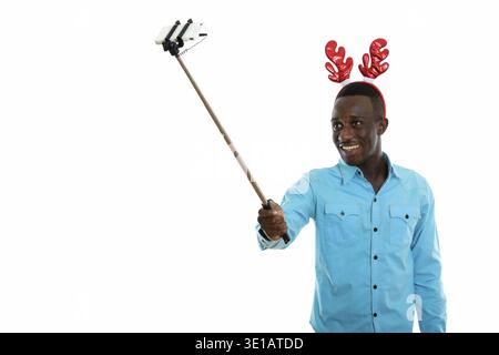 Studio shot der jungen schwarzen afrikanischen Mann glücklich lächelnd und Rentier Stirnband tragen, während der Einnahme von selfie Bild mit Handy auf selfie Stick Stockfoto