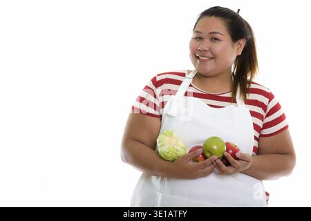 Studio shot der Jungen gerne Fett asiatische Frau lächelnd, während Sie Obst und Gemüse mit Schürze Stockfoto