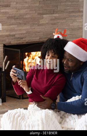 Afroamerikanisches Paar, das am Kamin sitzt, Selfie am Telefon macht, Weihnachtsmütze, Kuscheldecke Stockfoto