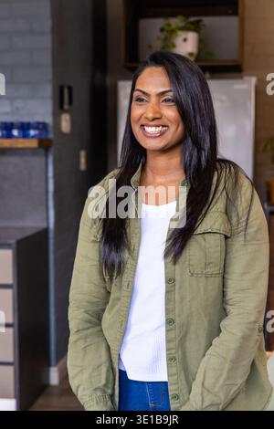 Indische Frau lächelt und steht in der Küche mit einem olivfarbenen Hemd in der Nähe einer blauen Brille und eines Kühlschranks Stockfoto