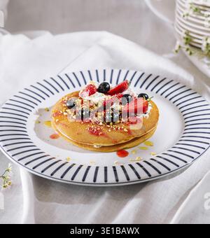 Köstlicher Stapel Pfannkuchen auf Teller mit blauen Streifen, belegt mit Erdbeeren, Heidelbeeren, Pistazien und Schlagsahne, getränkt in Sirup Stockfoto