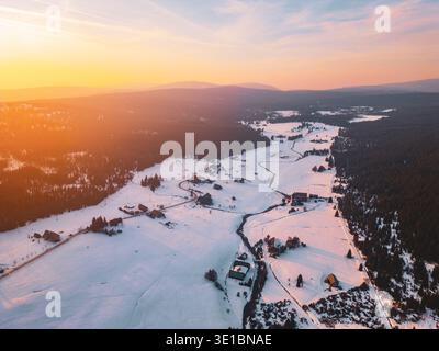 Aus der Vogelperspektive zeigt das Bergdorf Jizerka, das mit Schnee bedeckt ist. Die Landschaft bietet Häuser, Bäume und gewundene Pfade unter einem hellen Himmel bei Sonnenuntergang. Stockfoto