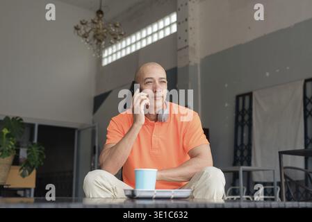 Hübscher Mann mittleren Alters in orangefarbenem Poloshirt am Kaffeetisch mit Kaffeetasse, Kopfhörern und Smartphone. Technologie und Lifestyle c Stockfoto