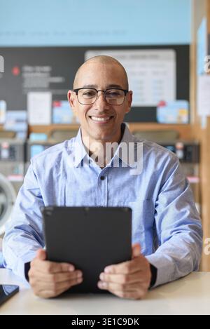 Glatzkopf in blauem Hemd und Brille mit Tablet-Computer, während er in einer modernen Wäscherei sitzt. Digitaler Lebensstil und echtes Geschäftskonzept Stockfoto