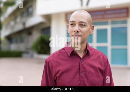 Stilvoller, kahlköpfiger italienischer Mann in burgunderfarbenem Button-Down-Hemd, der sich selbstbewusst vor dem modernen Universitätsgebäude an sonnigen Sommertagen posiert. Städtisch Stockfoto