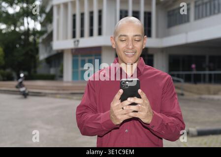 Glatzkopf italienischer Mann in burgunderrotem Hemd, der sein Smartphone überprüft, während er vor dem Universitätsgebäude bei hellem Sonnenlicht steht. Das Konzept des digitalen Kombi Stockfoto