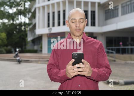 Glatzkopf italienischer Mann in burgunderrotem Hemd, der sein Smartphone überprüft, während er vor dem Universitätsgebäude bei hellem Sonnenlicht steht. Das Konzept des digitalen Kombi Stockfoto