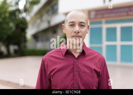 Stilvoller, kahlköpfiger italienischer Mann in burgunderfarbenem Button-Down-Hemd, der sich selbstbewusst vor dem modernen Universitätsgebäude an sonnigen Sommertagen posiert. Städtisch Stockfoto