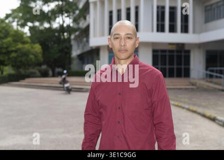 Stilvoller, kahlköpfiger italienischer Mann in burgunderfarbenem Button-Down-Hemd, der sich selbstbewusst vor dem modernen Universitätsgebäude an sonnigen Sommertagen posiert. Städtisch Stockfoto