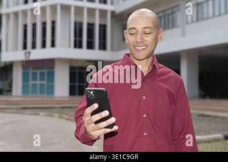 Glatzkopf italienischer Mann in burgunderrotem Hemd, der sein Smartphone überprüft, während er vor dem Universitätsgebäude bei hellem Sonnenlicht steht. Das Konzept des digitalen Kombi Stockfoto