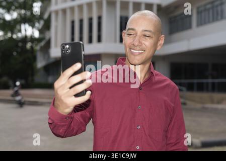 Glatzkopf italienischer Mann in burgunderrotem Hemd, der sein Smartphone überprüft, während er vor dem Universitätsgebäude bei hellem Sonnenlicht steht. Das Konzept des digitalen Kombi Stockfoto