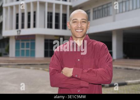 Stilvoller, kahlköpfiger italienischer Mann in burgunderfarbenem Button-Down-Hemd, der sich selbstbewusst vor dem modernen Universitätsgebäude an sonnigen Sommertagen posiert. Städtisch Stockfoto