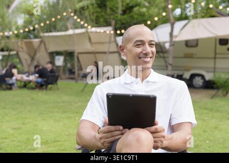 Stilvoller kahlköpfiger Mann in einem weißen Poloshirt, der sich auf einem Campingplatz neben einem Wohnmobil mit einem Tablet-Computer entspannt. Outdoor-Reise- und Camping-Lifestyle-Konzept Stockfoto