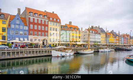 Besucher schlendern entlang der lebendigen Kanäle von Nyhavn, bewundern die historischen Gebäude und genießen lokale Cafés. Boote schweben sanft im Wasser unter einem bewölkten Himmel. Stockfoto