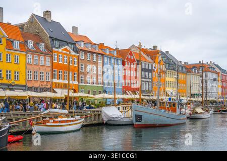 Touristen schlendern entlang des lebendigen Nyhavn-Kanals in Kopenhagen und bewundern die farbenfrohen historischen Gebäude und Boote, die auf dem Wasser verankert sind. Die Atmosphäre ist lebhaft, trotz des grauen Himmels. Stockfoto